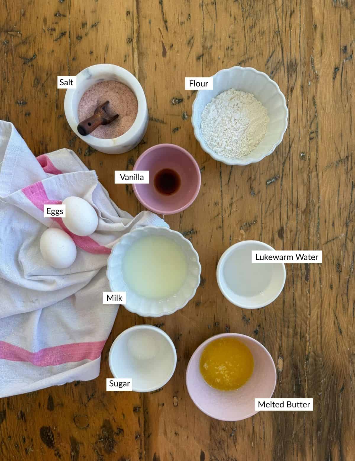 Sweet crepe recipe ingredients arranged on a wooden surface including eggs, flour, vanilla extract, sugar, melted butter, salt, and milk.