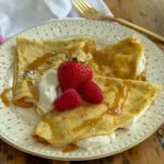 Two sweet crepes filled with whipped cream and folded in triangles on a vintage star-patterned plate, topped with fresh strawberries, raspberries, and caramel sauce.