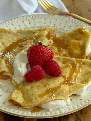 Two sweet crepes filled with whipped cream and folded in triangles on a vintage star-patterned plate, topped with fresh strawberries, raspberries, and caramel sauce.