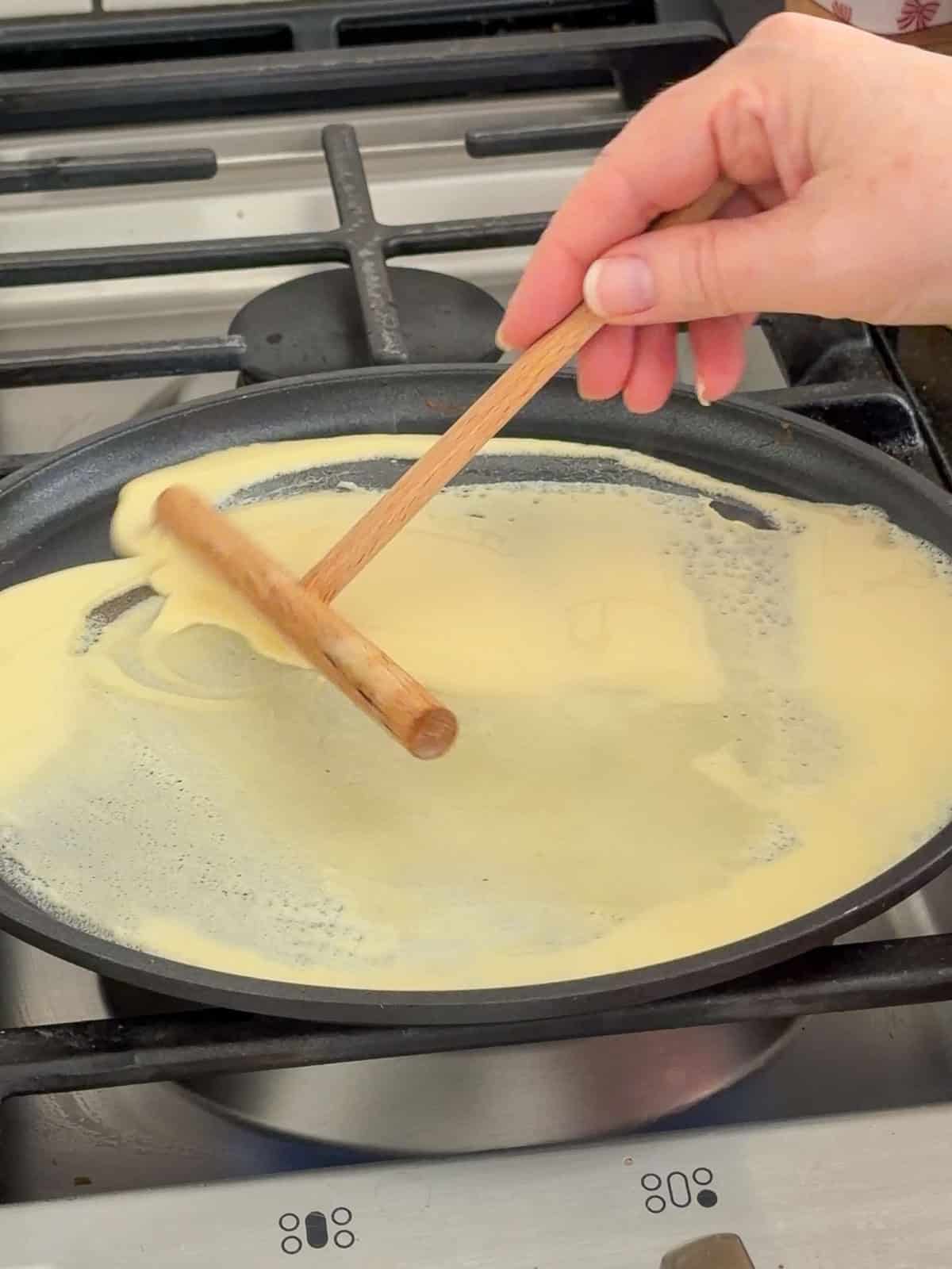 Sweet crepe batter being spread in a circular motion with a wooden T-spreader in a large nonstick pan on a gas stovetop.