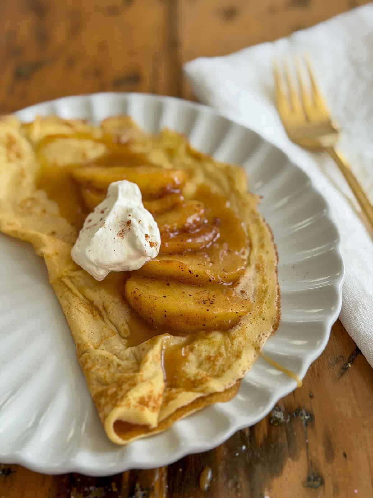 Sweet crepe filled with cinnamon saut&eacute;ed apple slices and topped with a dollop of whipped cream on a white scalloped plate.