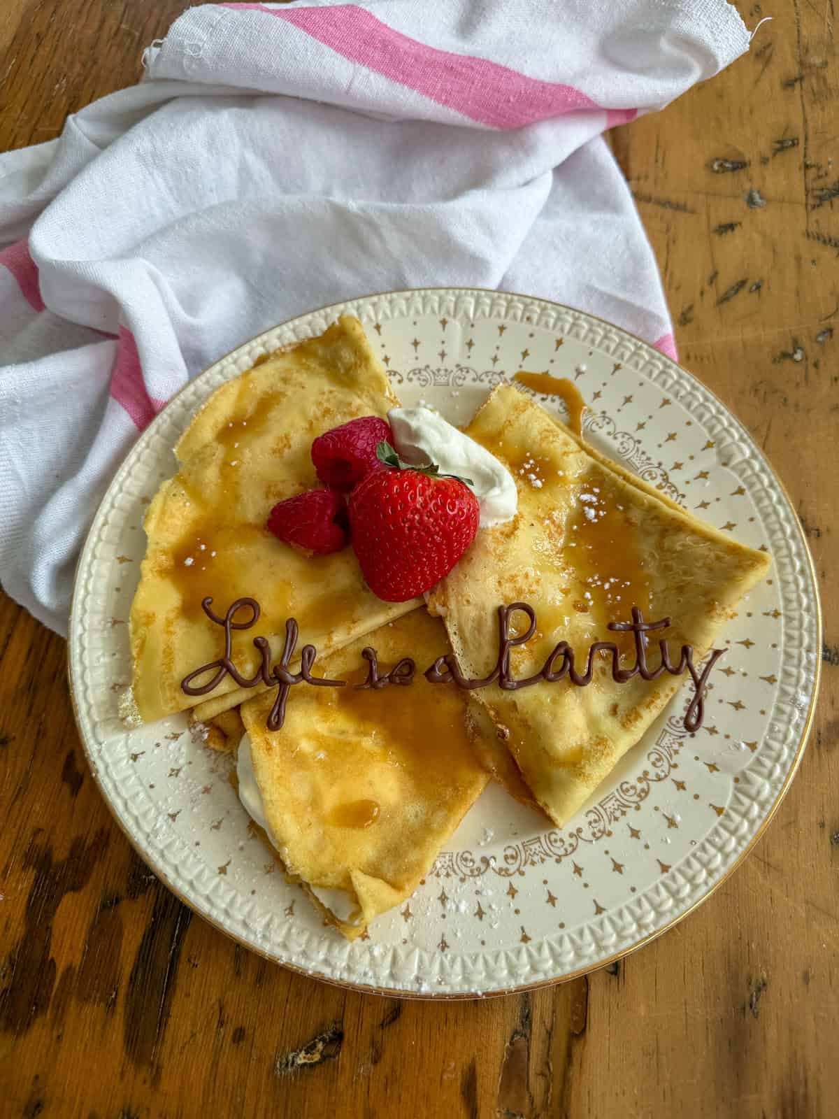 Sweet crepes on a vintage plate with whipped cream, fresh strawberries, raspberries, caramel sauce drizzle, and "Life is a Party" written in chocolate.