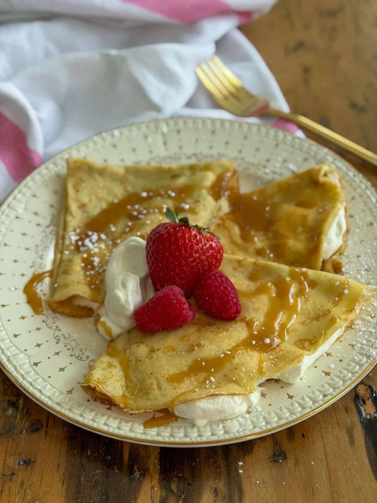 Sweet crepes filled with whipped cream and folded in triangles on a vintage plate, topped with fresh strawberries, raspberries, powdered sugar, and a caramel sauce drizzle.