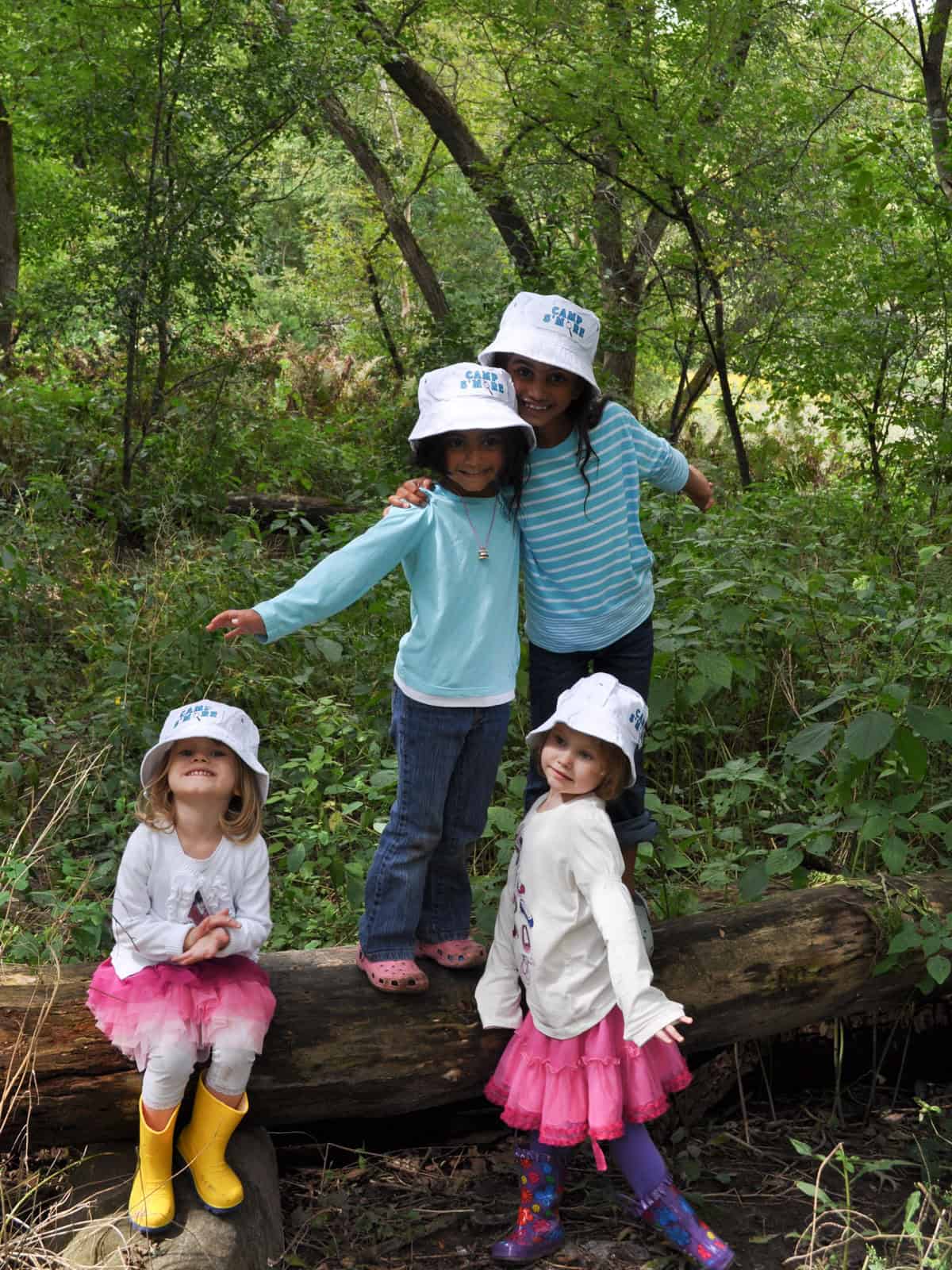 Four children in white Camp S'more bucket hats posing together on a fallen log.