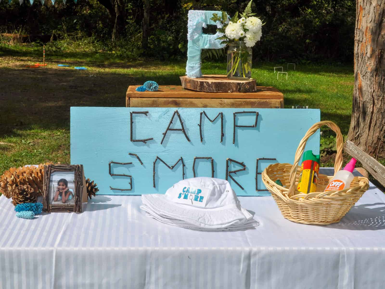 Camp S'more welcome table with a twig lettered sign, wrapped letter P, camp hats, and a bug spray basket.