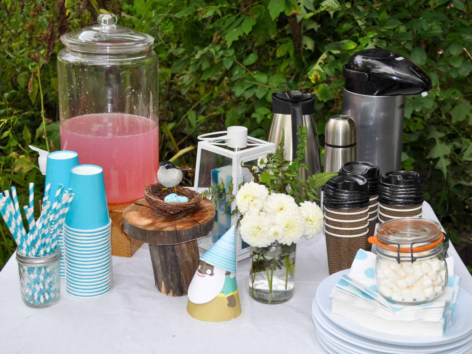 Camping party drink station with pink lemonade, paper straws, and coffee thermoses.
