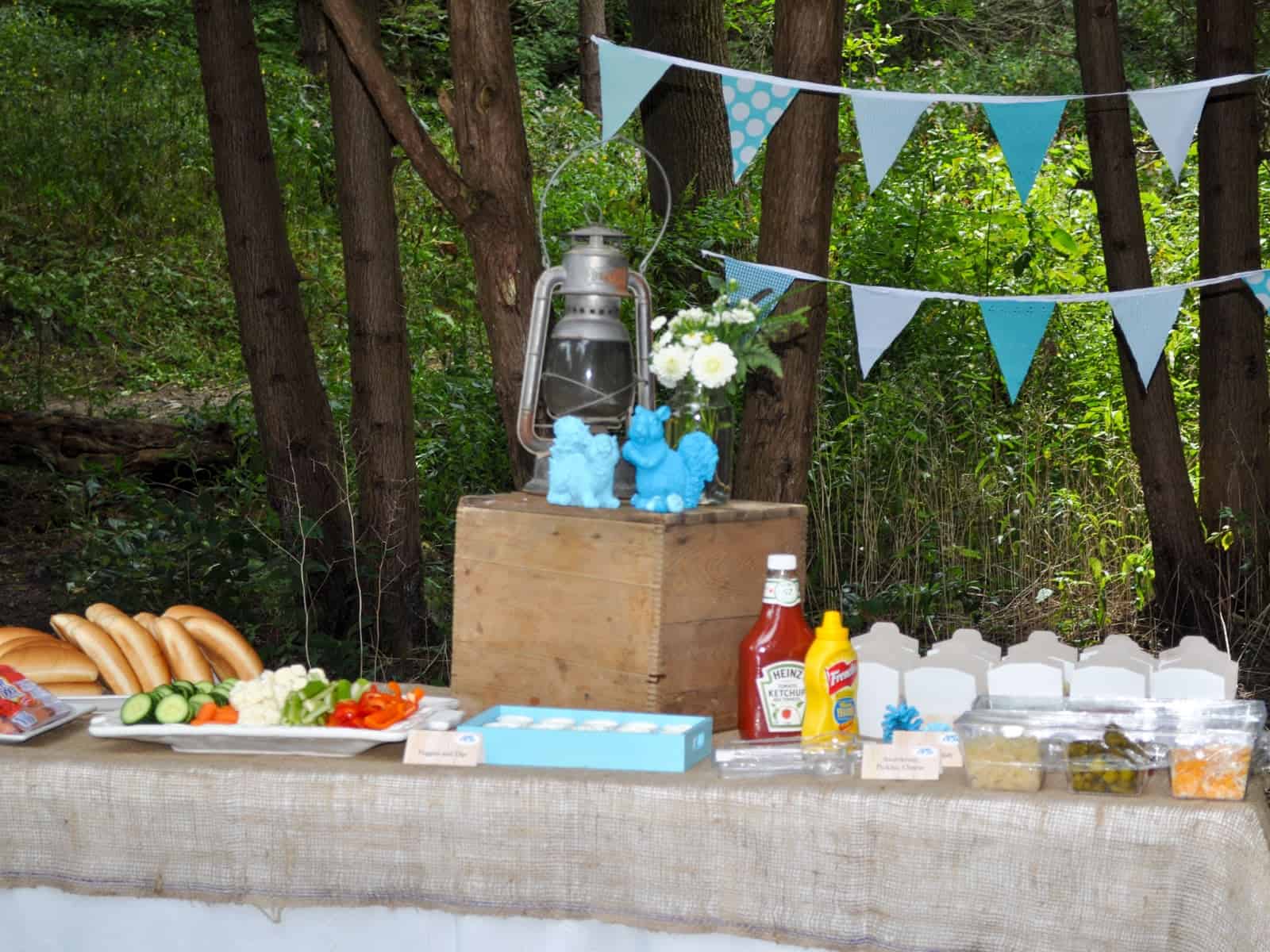 Hot dog buffet with buns, chopped vegetables, ketchup, and mustard on a burlap covered table.