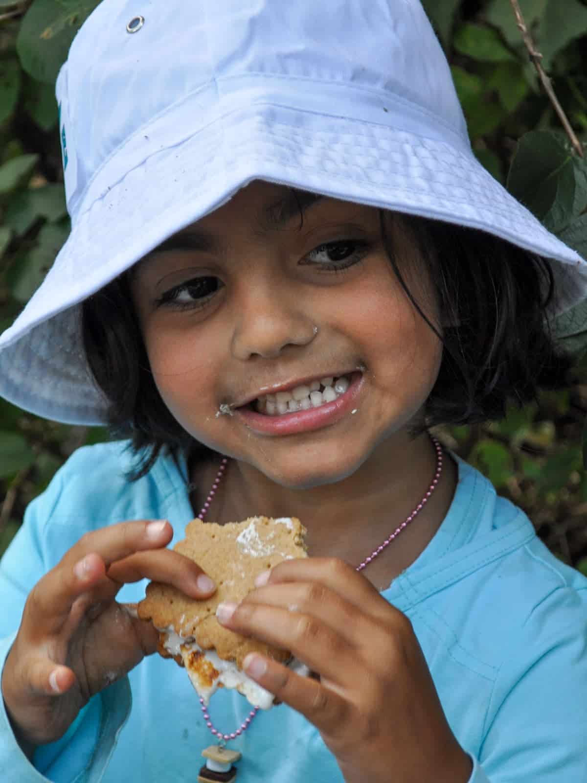 Girl in a white bucket hat smiling with a marshmallow smeared s'more.