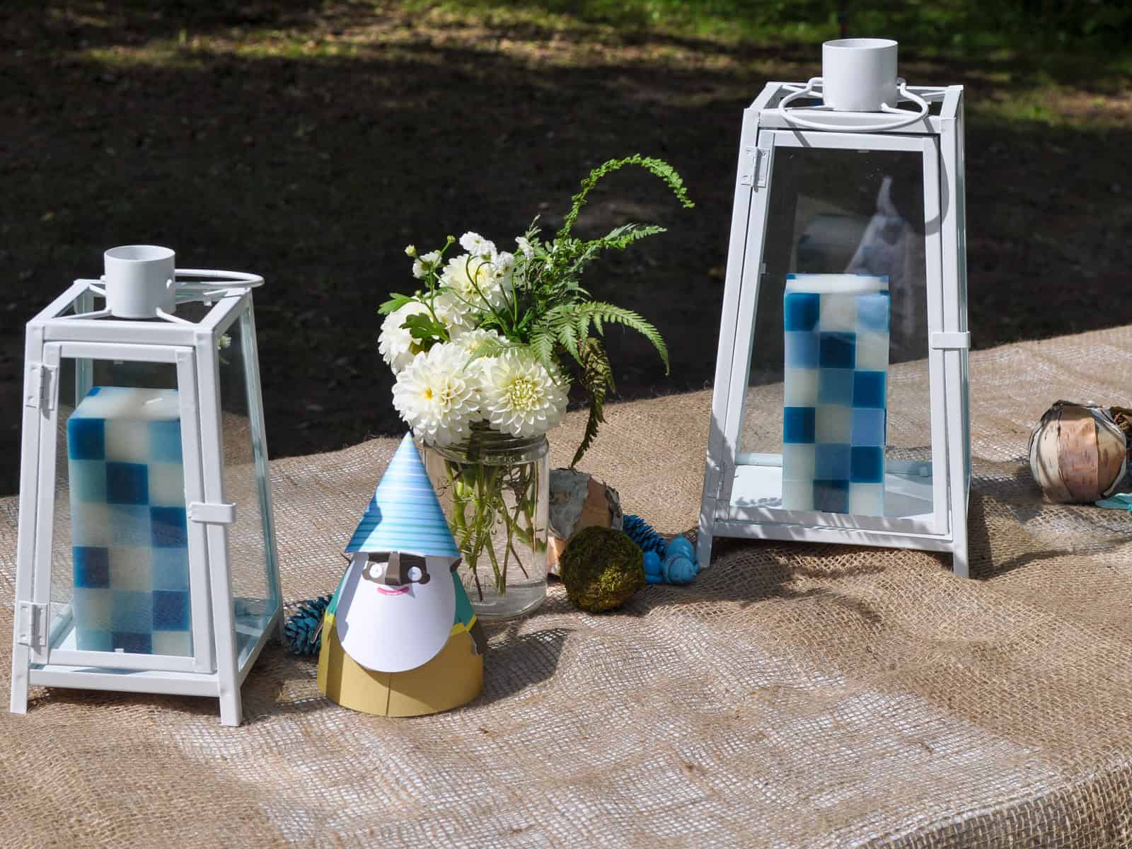 White lanterns with checkered candles flanking a paper gnome party hat and white dahlias.