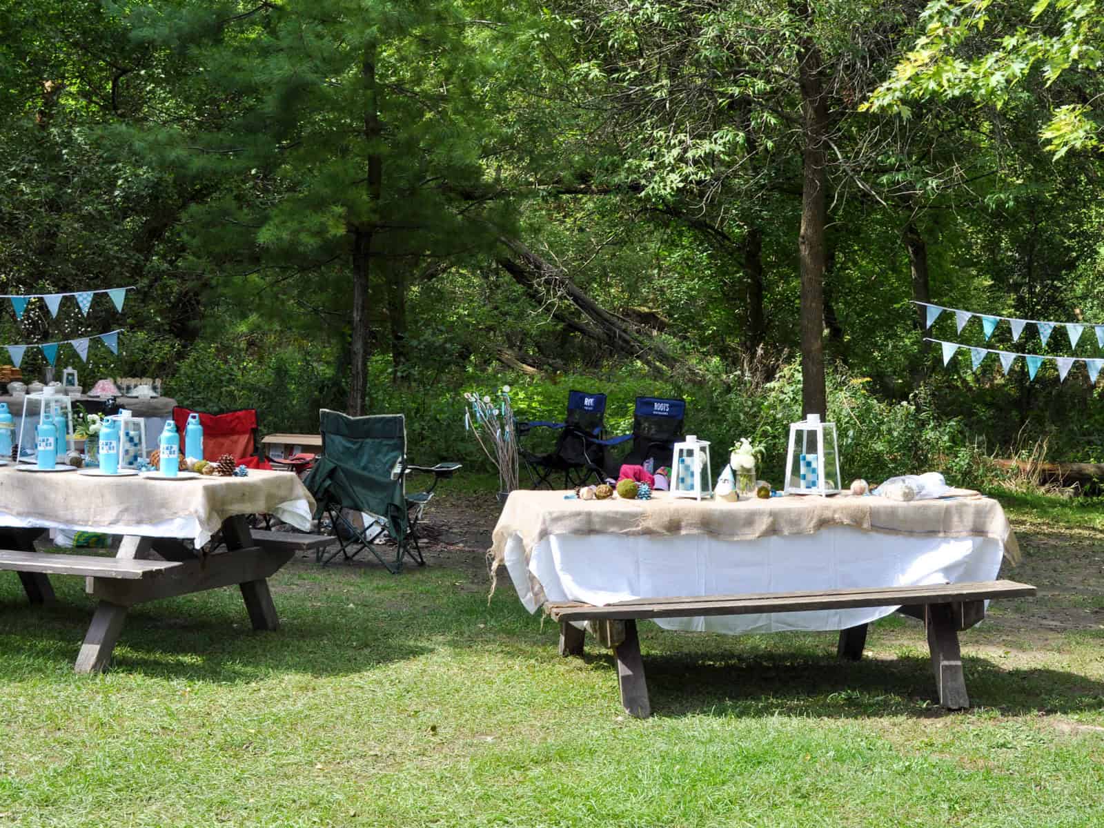 Two picnic tables set for a camping birthday party with aqua bunting strung between the trees.