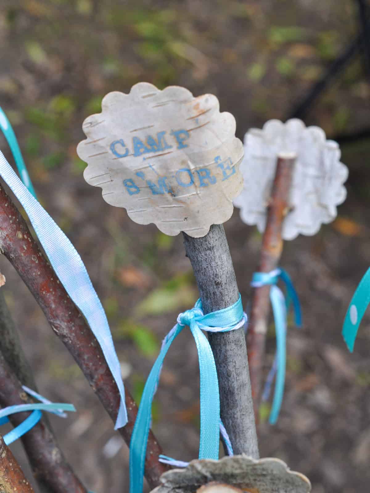 Birch bark tag stamped with Camp S'more tied to a roasting stick with aqua ribbon.