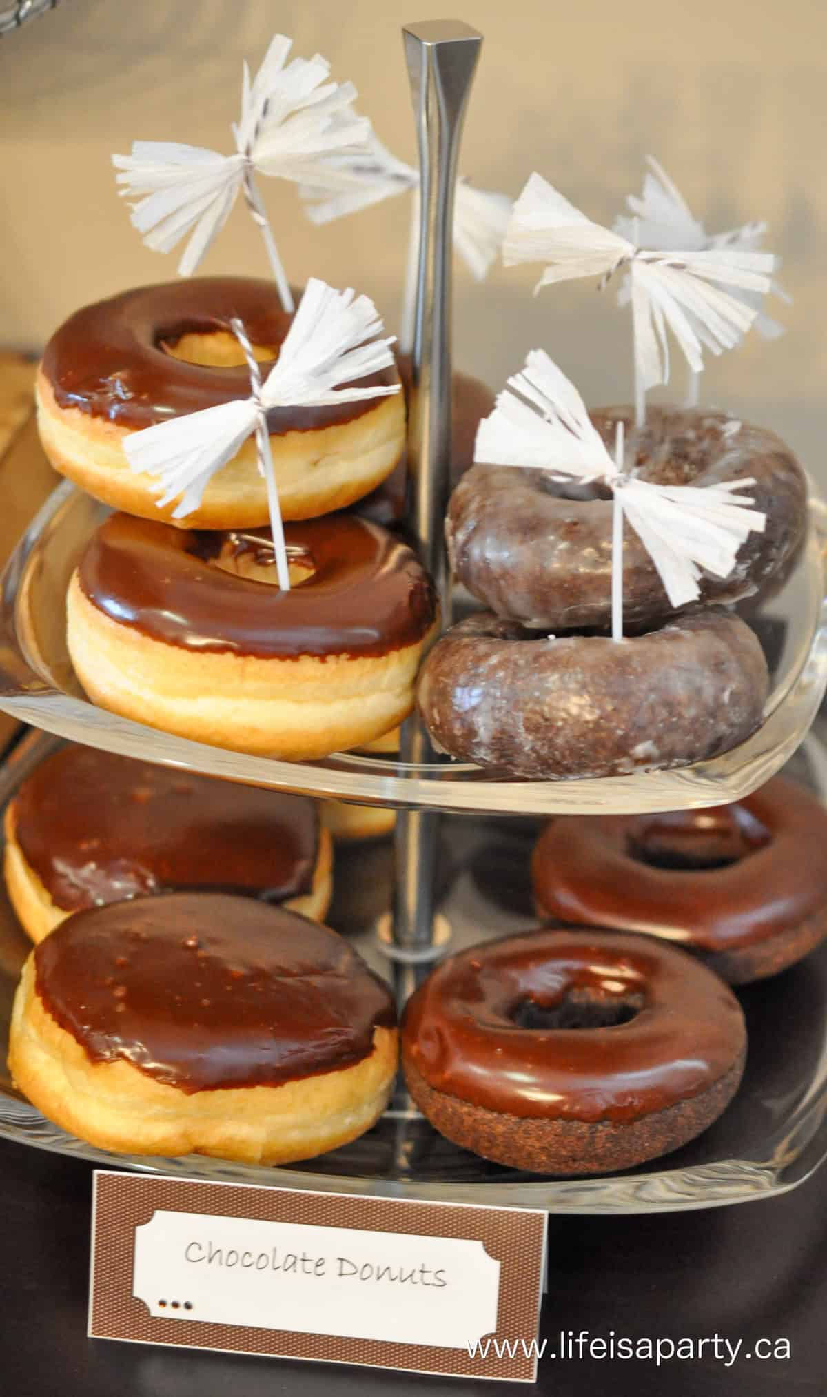 Assorted chocolate donuts on a glass tiered serving tray on a chocolate themed dessert table.