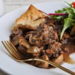 Steak Guinness and mushroom pie served on a white plate with golden puff pastry, tender beef, mushrooms in rich gravy, and fresh salad.