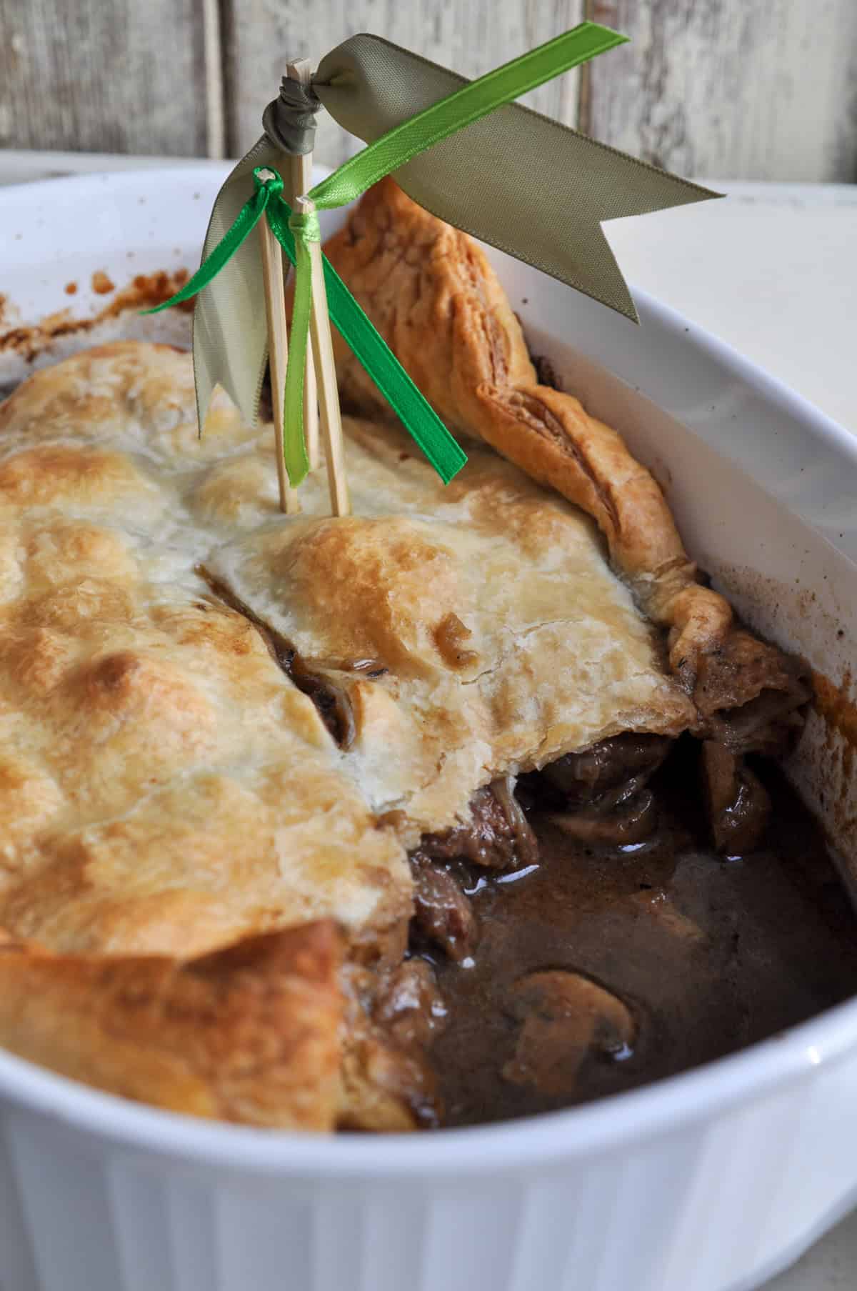 Steak Guinness and mushroom pie in white ceramic baking dish with golden puff pastry crust and decorative toothpick flags.