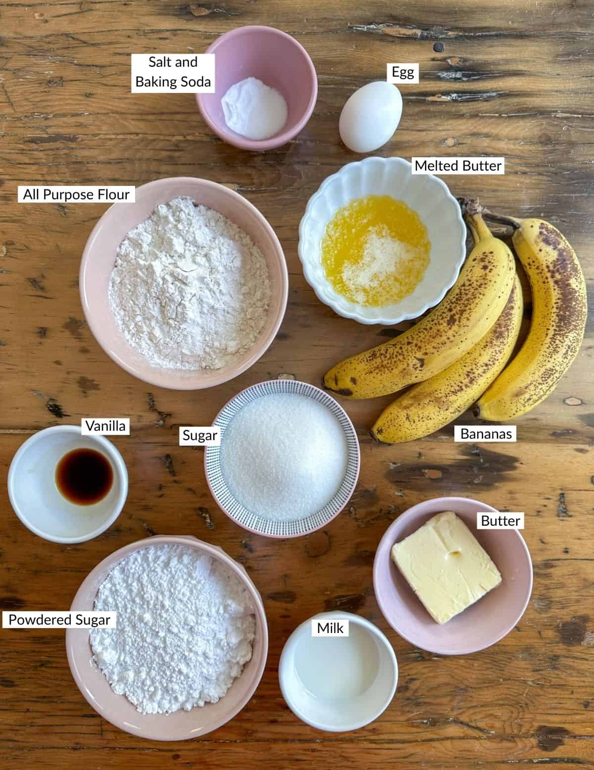 Banana snack cake ingredients in small bowls on a wood table.