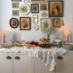 Crepe bar on white sideboard with vintage floral gallery wall, lace tablecloth, tiered stand with vanilla and chocolate crepes, fresh fruit, and assorted toppings in mismatched vintage china.