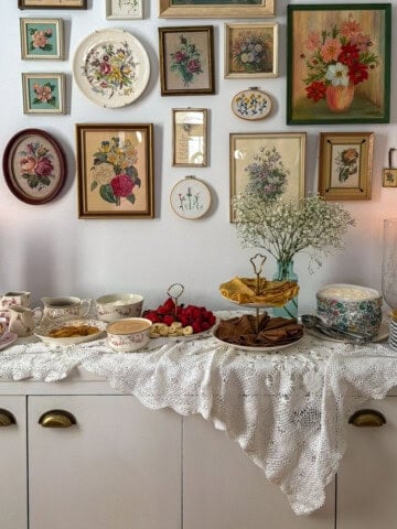Crepe bar on white sideboard with vintage floral gallery wall, lace tablecloth, tiered stand with vanilla and chocolate crepes, fresh fruit, and assorted toppings in mismatched vintage china.
