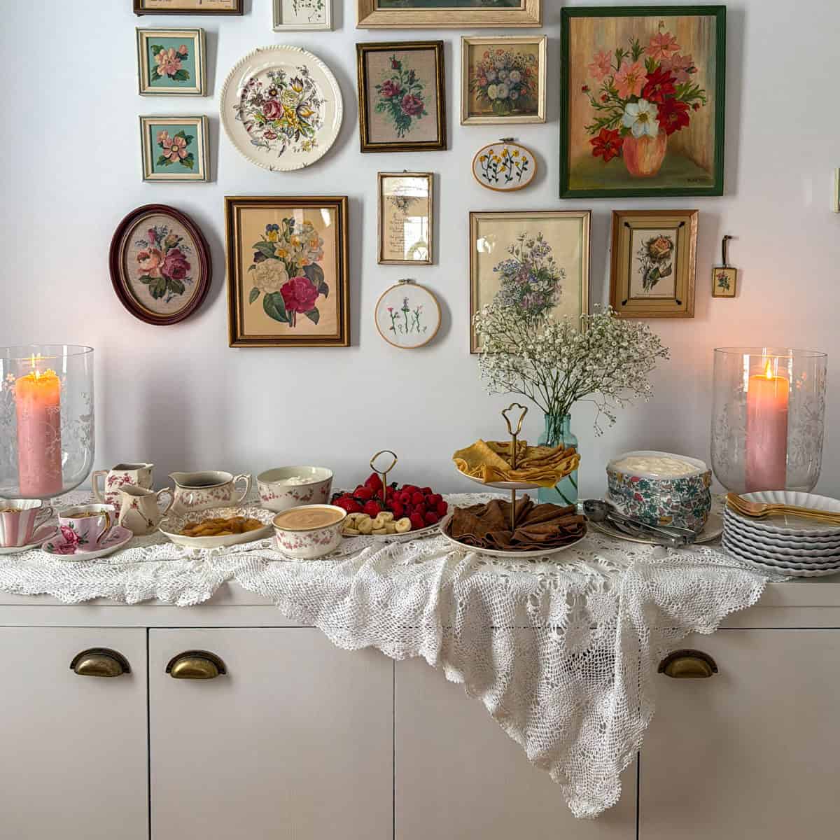 Crepe bar on white sideboard with vintage floral gallery wall, lace tablecloth, tiered stand with vanilla and chocolate crepes, fresh fruit, and assorted toppings in mismatched vintage china.