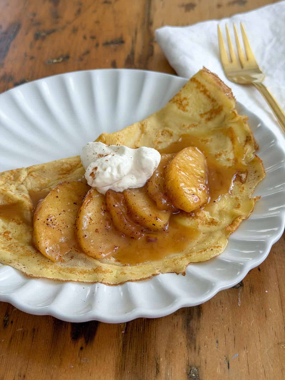 Vanilla crepe folded on a white scalloped plate and topped with sauteed cinnamon apples, caramel sauce, and whipped cream with a gold fork alongside.