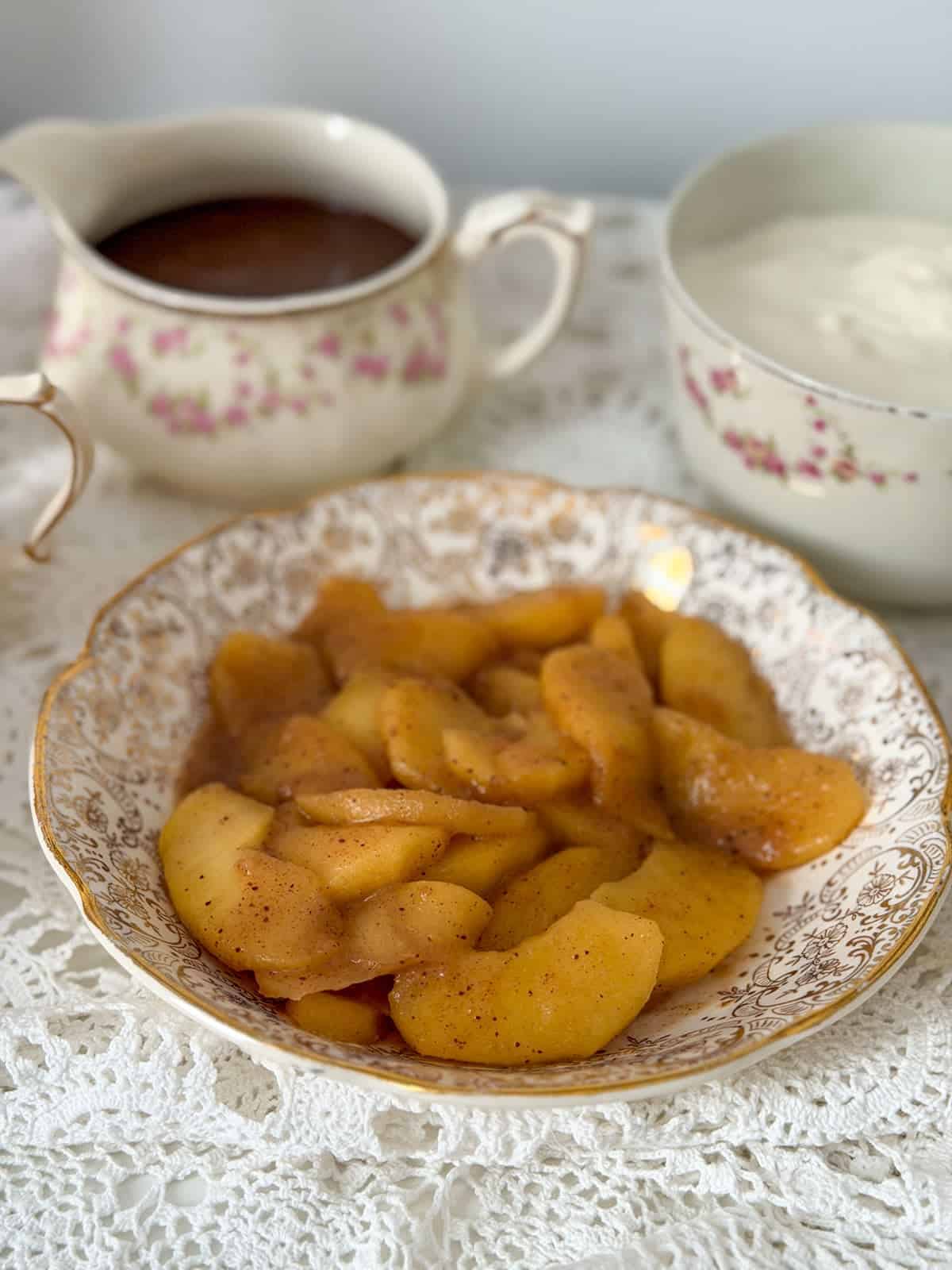 Warm cinnamon sauteed apple slices in a gold-trimmed vintage china plate with a chocolate sauce pitcher and whipped cream bowl in the background.