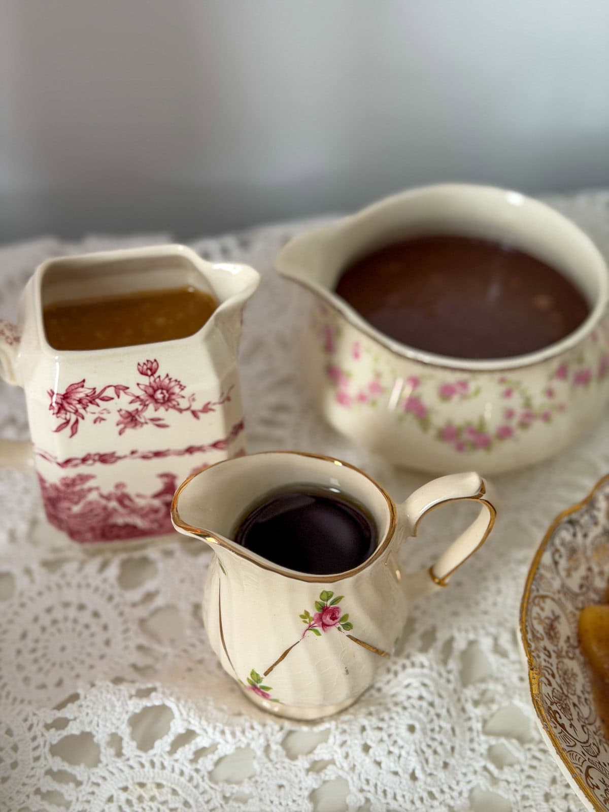 Three mismatched vintage china pitchers holding caramel sauce, hot fudge sauce, and maple syrup on a lace tablecloth.