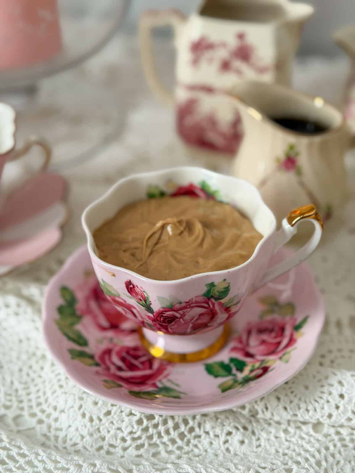 Hazelnut cream served in a pink rose vintage teacup and saucer on a lace tablecloth with china sauce pitchers in the background.