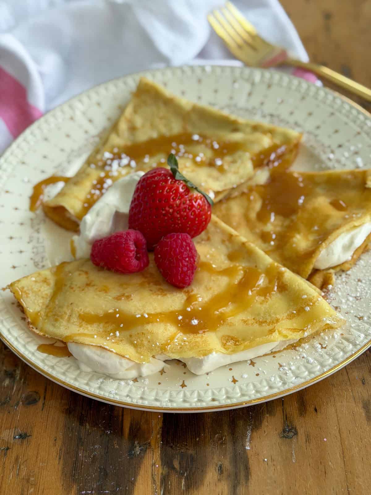 Two vanilla crepes folded into quarters on a cream and gold plate, filled with whipped cream and topped with caramel sauce, a strawberry, and raspberries with a gold fork alongside.