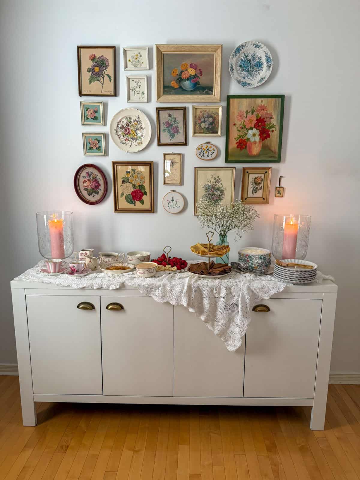 Full crepe bar setup on white sideboard with vintage floral gallery wall, lace tablecloth, tiered crepe stand, and toppings in mismatched china with pink pillar candles.