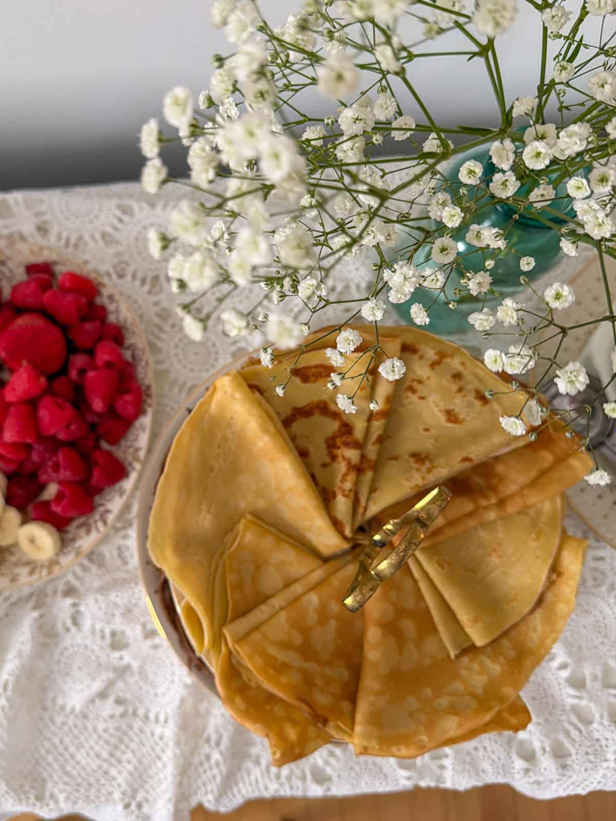 Vanilla crepes folded into triangles and fanned on a round plate with fresh raspberries and baby's breath flowers alongside.