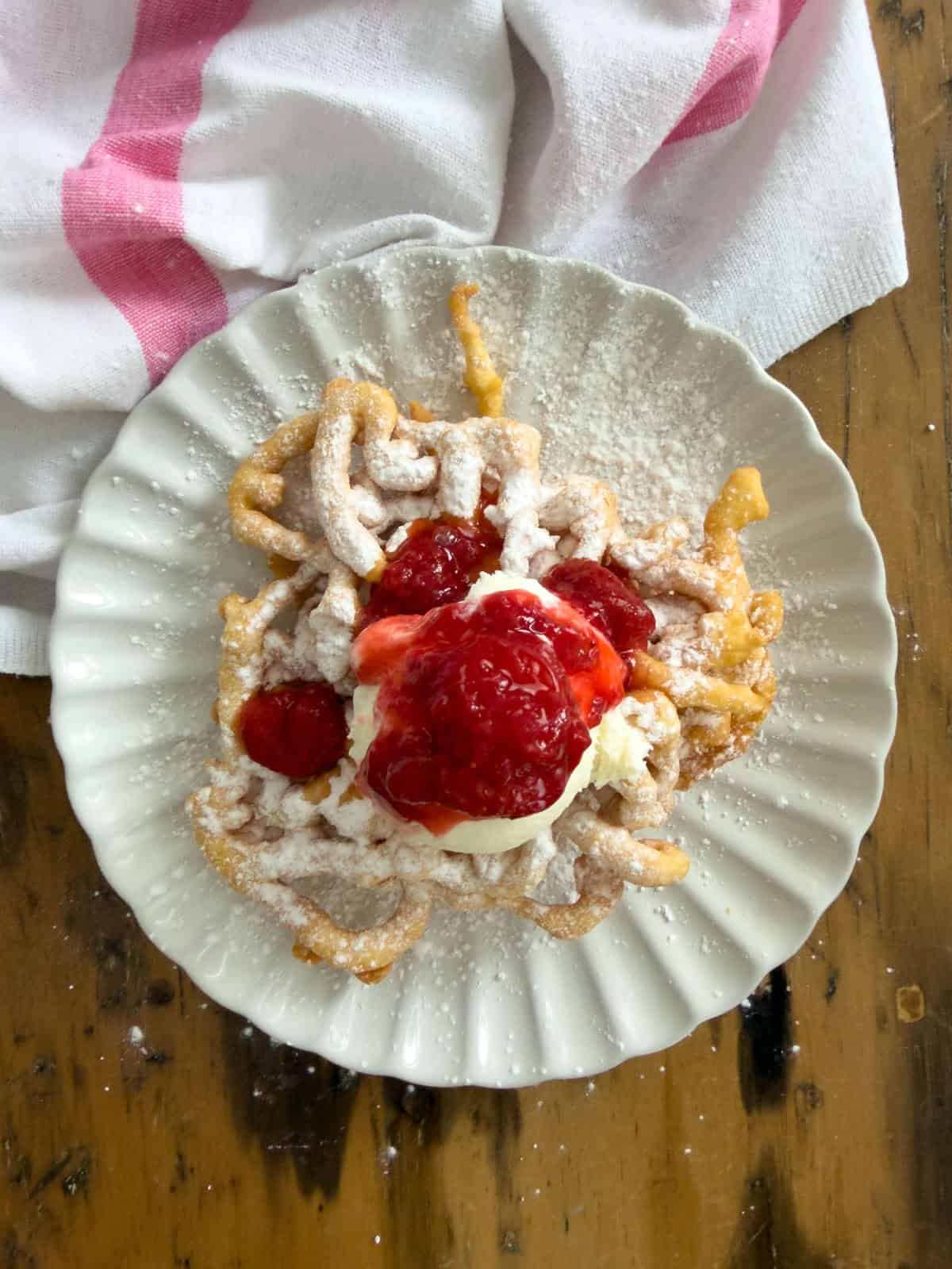 Homemade funnel cake with strawberry compote, vanilla ice cream, and powdered sugar, on a white plate.