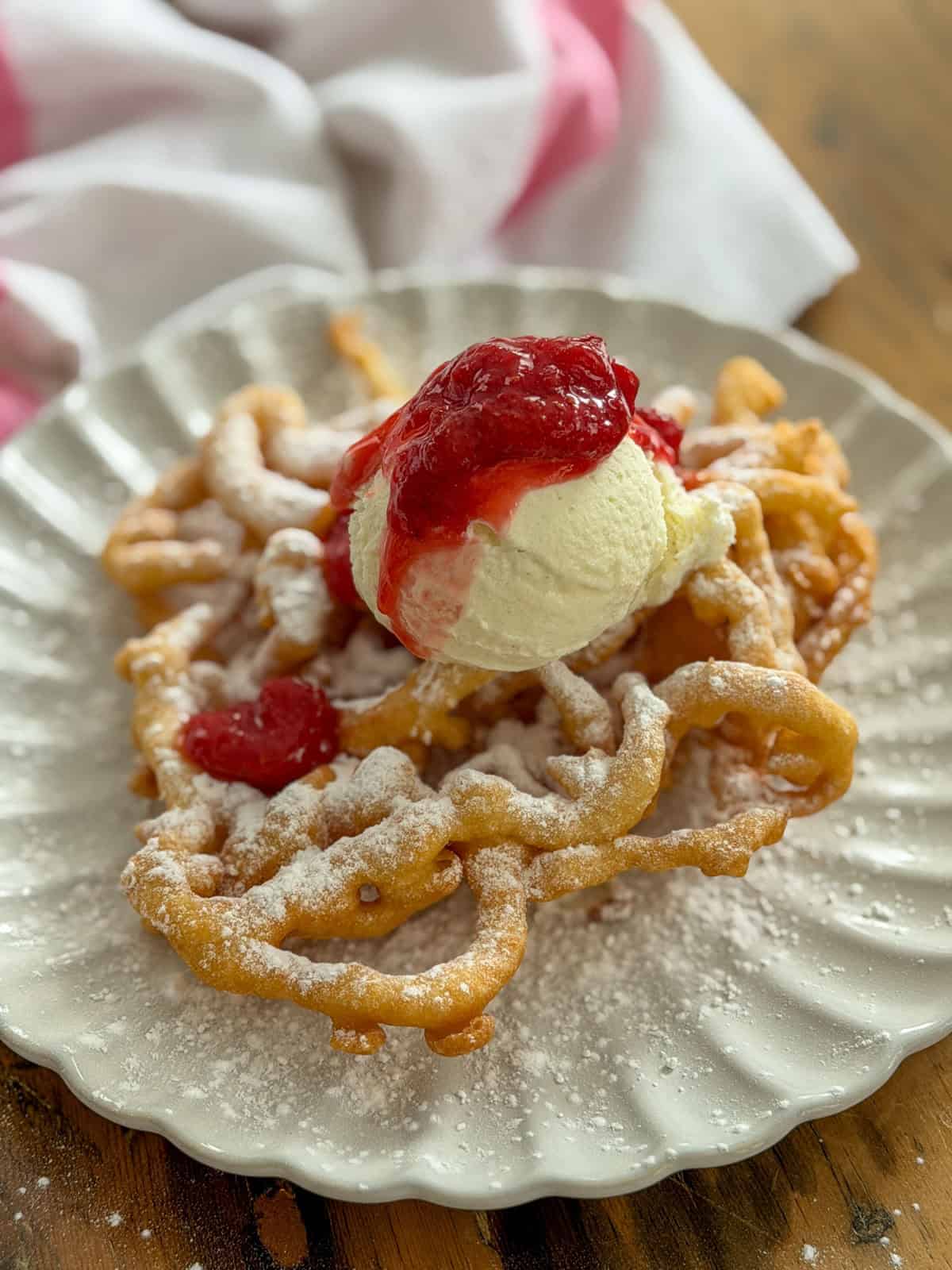 Fair funnel cake with strawberry compote and vanilla ice cream dusted with powdered sugar on a white scalloped plate.