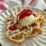 Fair funnel cake with strawberry compote and vanilla ice cream dusted with powdered sugar on a white scalloped plate.