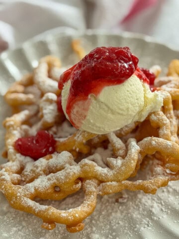 Fair funnel cake with strawberry compote and vanilla ice cream dusted with powdered sugar on a white scalloped plate.