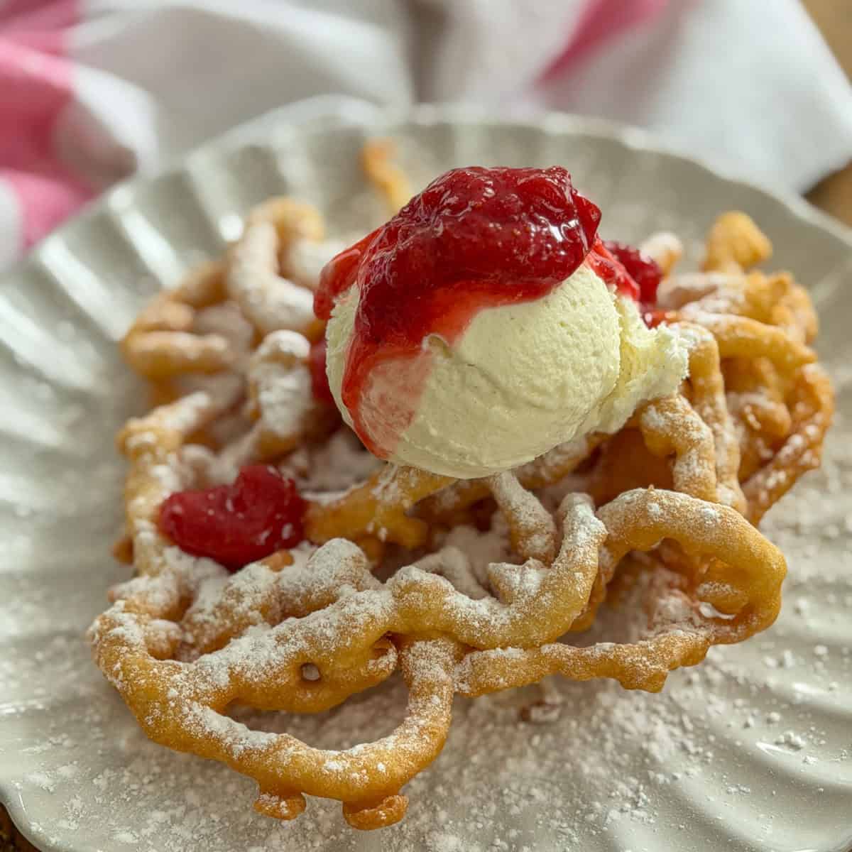 Fair funnel cake with strawberry compote and vanilla ice cream dusted with powdered sugar on a white scalloped plate.