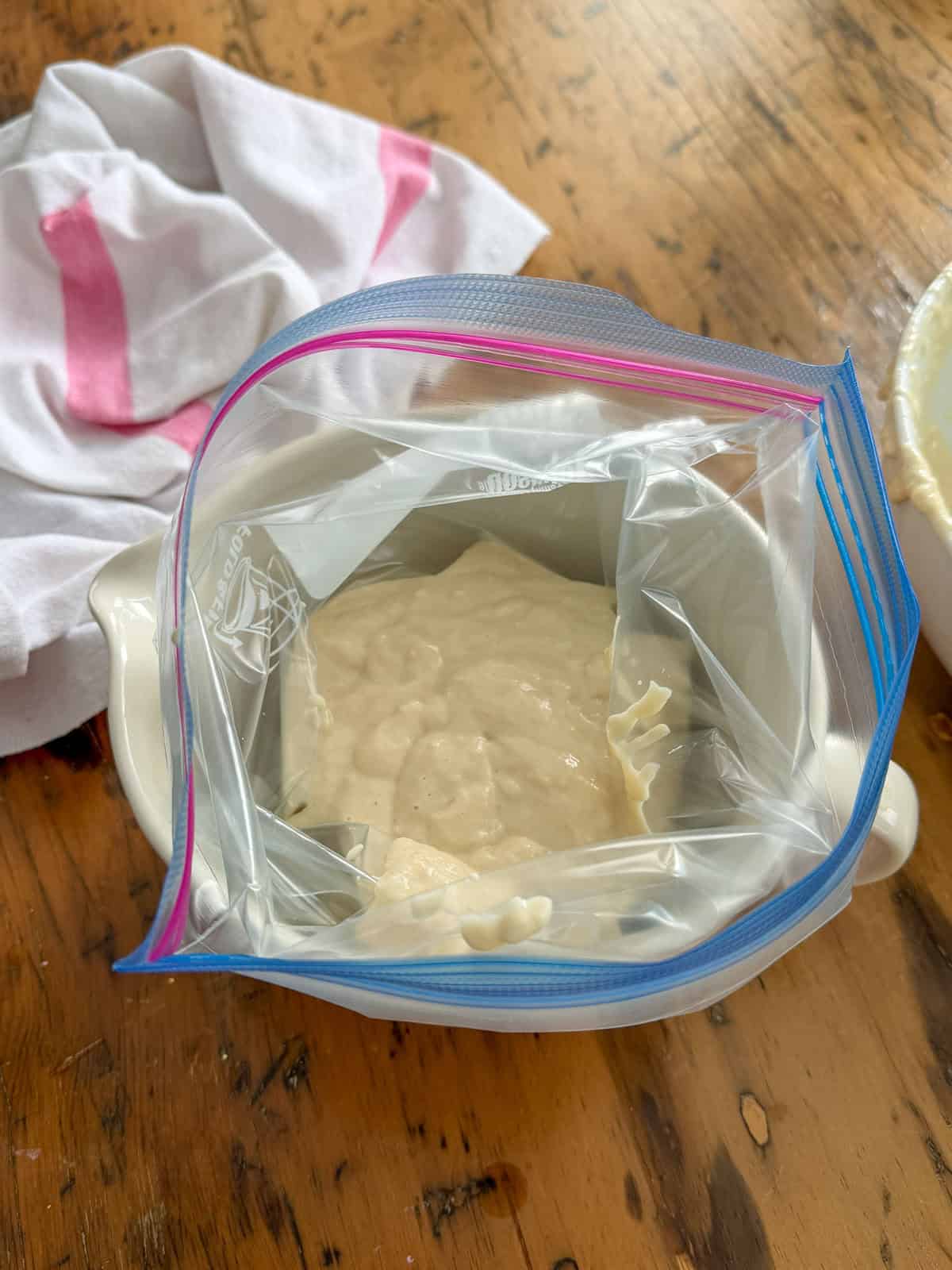 Funnel cake batter being poured into a ziplock freezer bag set inside a white bowl.