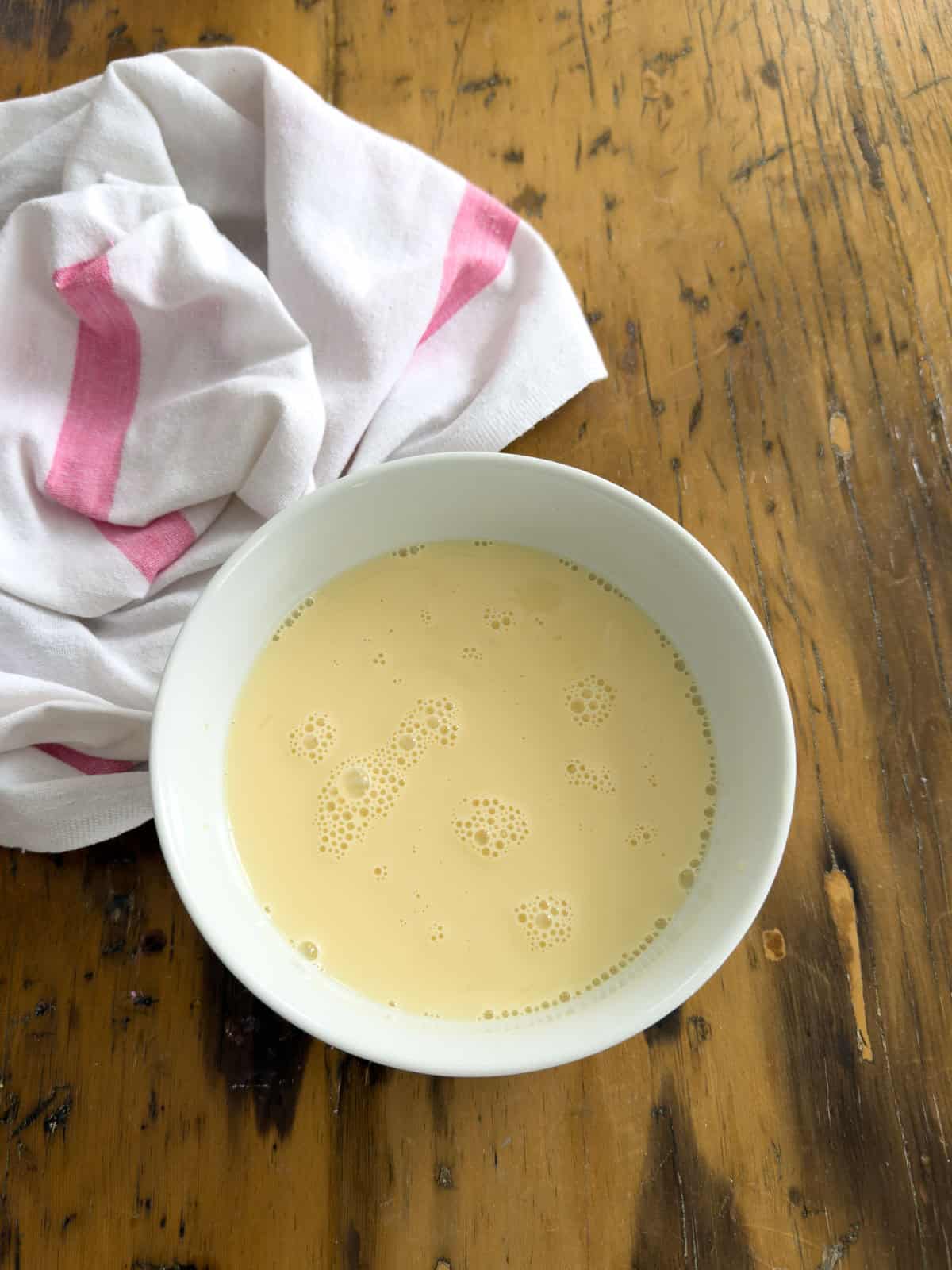 Eggs, milk, and vanilla whisked together in a white bowl on a wooden table.