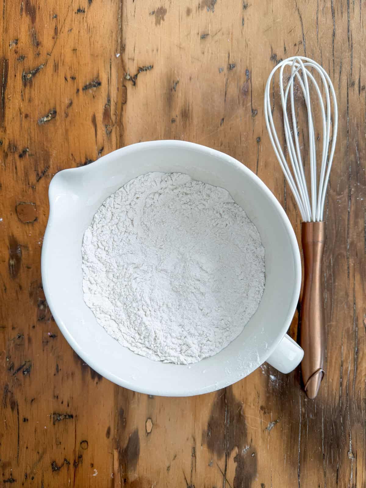 Sifted flour, baking powder, and salt combined in a white mixing bowl with a copper-handled whisk on a wooden surface.