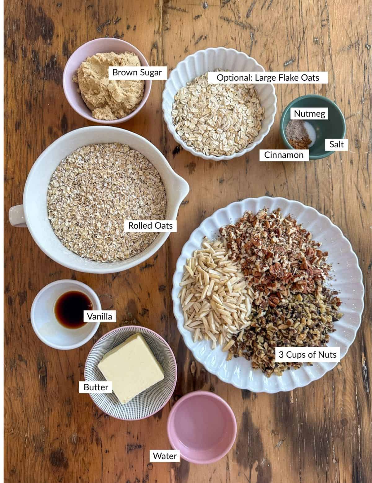 Crunchy granola ingredients laid out on wooden table including rolled oats, brown sugar, butter, vanilla, cinnamon, nutmeg, salt and mixed nuts.