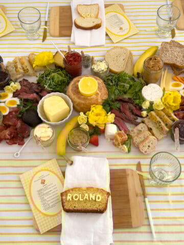 Butter themed toast party dining table set with yellow striped tablecloth, compound butter grazing runner down the center, baguette candle holders, and toasters at each end, white hutch and crystal chandelier in background.