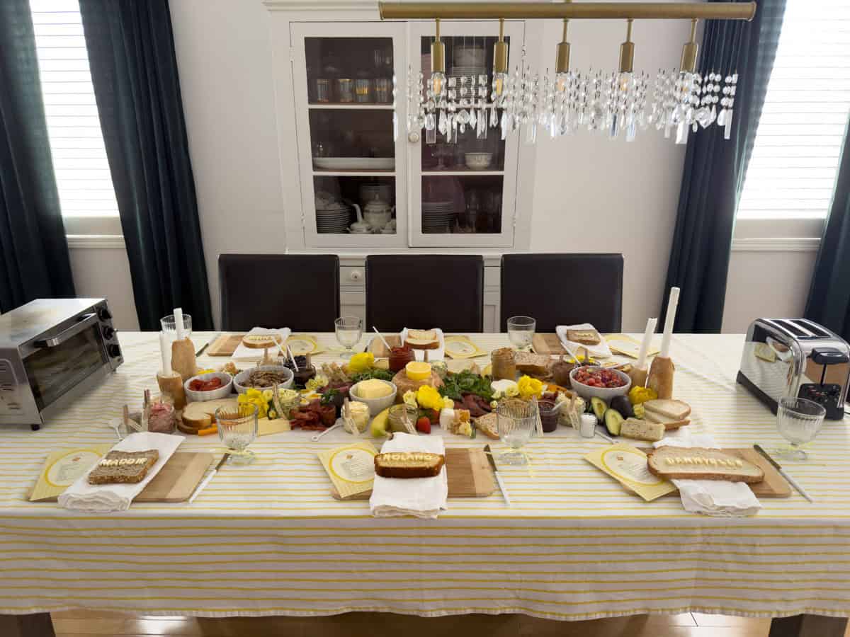 Butter themed toast party dining table set with yellow striped tablecloth showing toaster oven at the left end and chrome toaster at the right end flanking the full compound butter grazing spread with name bread place settings, bamboo cutting boards, and crystal chandelier above.