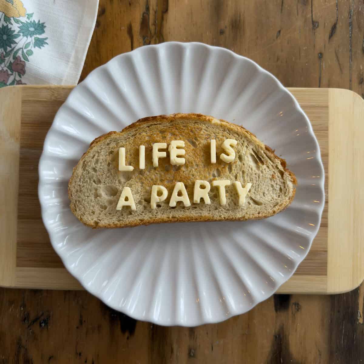 Slice of toasted sourdough bread on a white scalloped plate spelling out LIFE IS A PARTY in butter letter tiles on a bamboo cutting board.