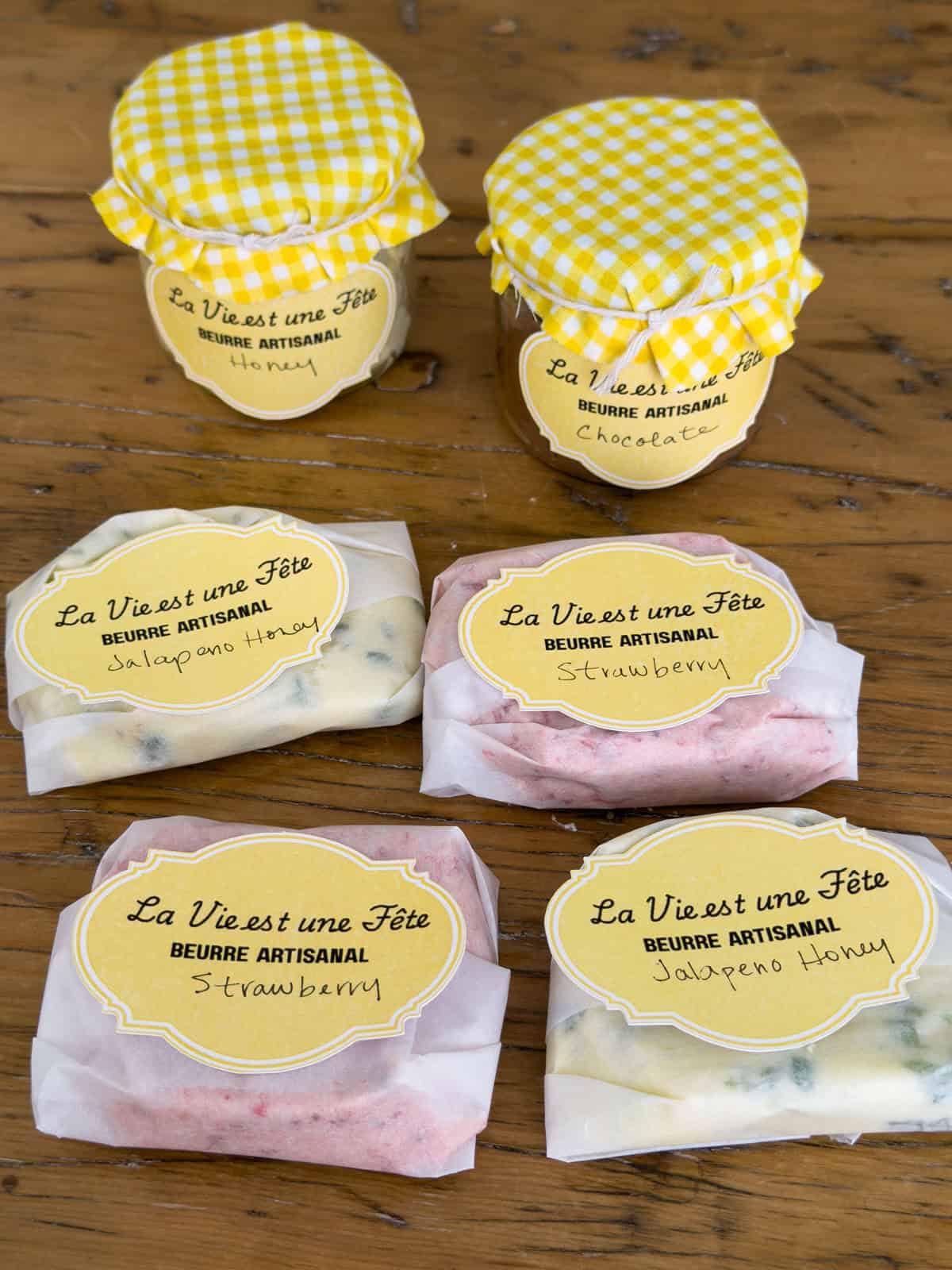 Compound butter party favors including two mason jars with yellow gingham fabric lids labeled Honey and Chocolate, and four parchment-wrapped butter portions labeled Jalapeno Honey and Strawberry with La Vie est une Fete Beurre Artisanal labels.