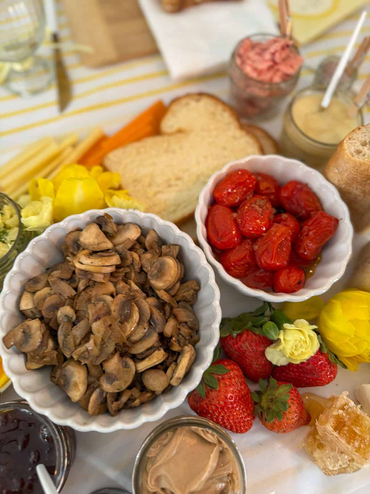 Overhead close up of white scalloped bowl of sauteed mushrooms and white scalloped bowl of roasted cherry tomatoes on a white parchment grazing surface with fresh strawberries, yellow roses and tulips, honeycomb, peanut butter jar, strawberry butter jar, and sliced bread visible around them.