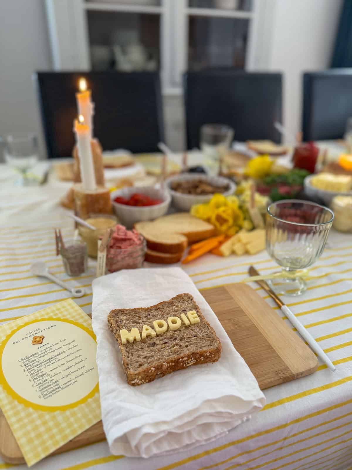 Slice of whole grain bread with butter letter tiles spelling MADDIE used as a name card place setting on a bamboo cutting board with lit taper candles in baguette holders, strawberry butter jar, and compound butter grazing spread glowing in candlelight in background.