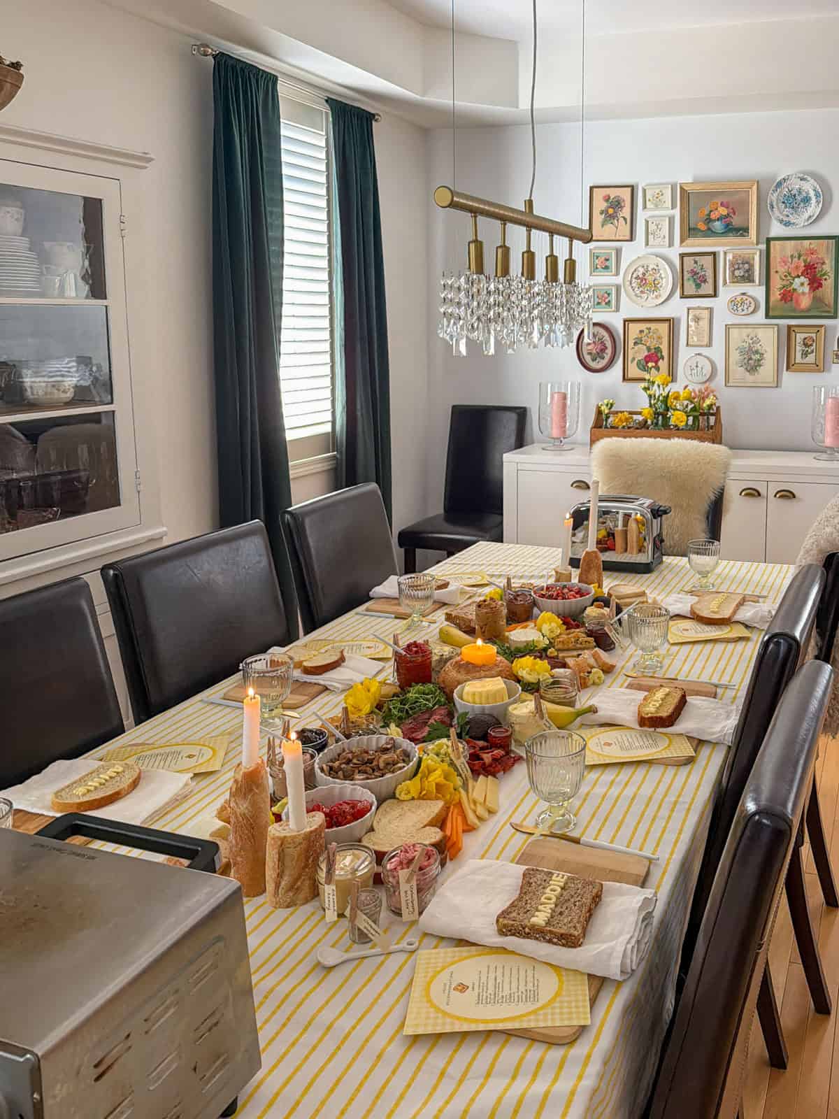 Dining room set for a butter themed toast party with yellow striped tablecloth, lit candles, compound butter grazing board, and gallery wall of botanical prints in background.