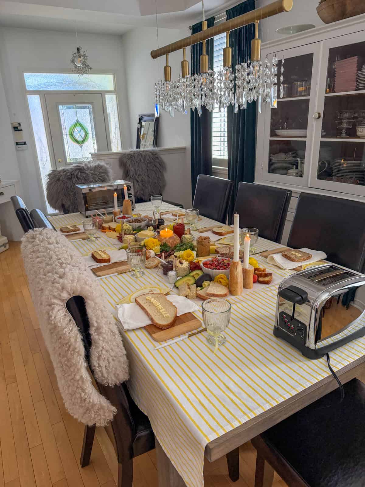 Butter toast party dining table from the side showing place settings with name breads, baguette candle holders, lit taper candles, and yellow flower arrangements along the grazing runner.