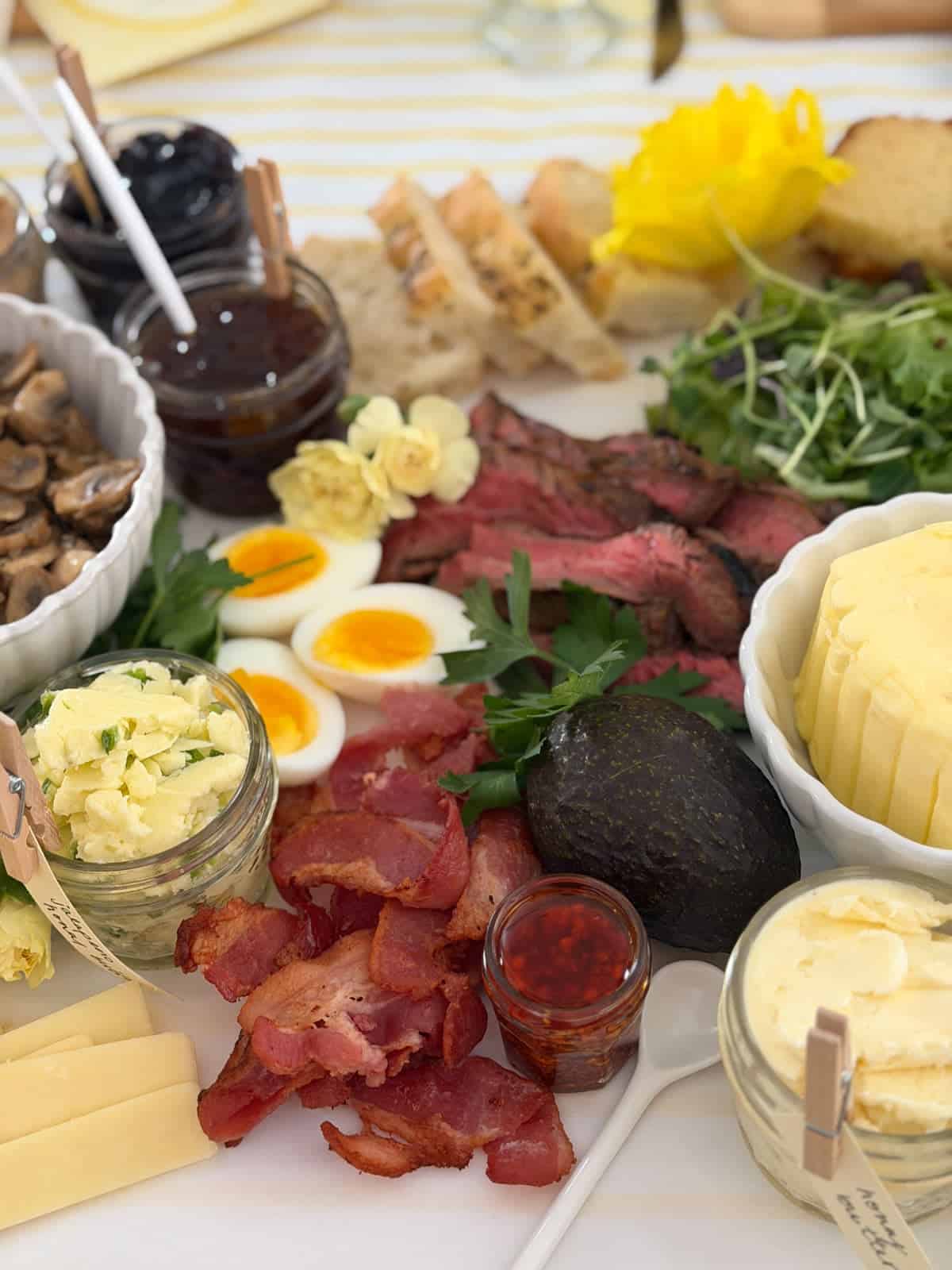 Overhead view of compound butter toast party grazing board with sliced flank steak, sauteed mushrooms, caramelized onion jam, blueberry vanilla jam, hazelnut creme, roasted cherry tomatoes, hard boiled eggs, fresh strawberries, and honeycomb.