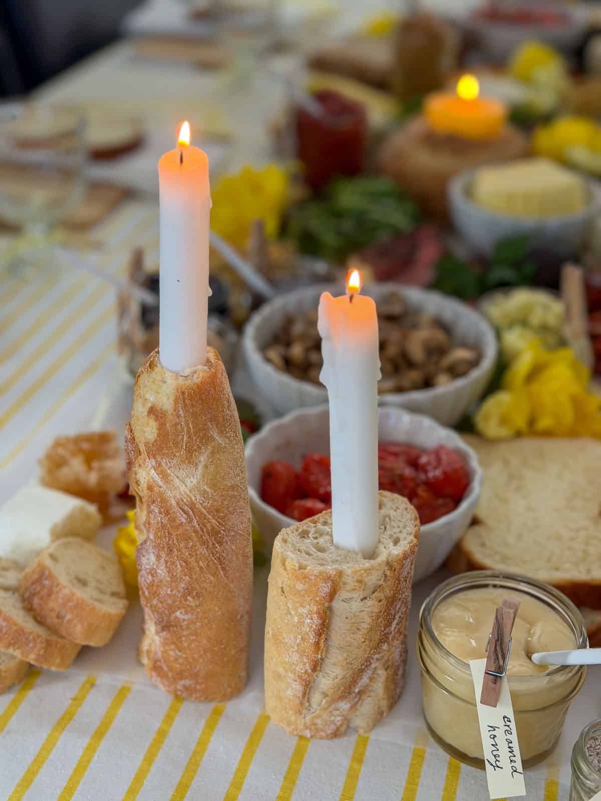 Two baguette sections used as taper candle holders on a white parchment grazing runner with roasted cherry tomatoes, sauteed mushrooms, strawberries, and honeycomb visible behind.