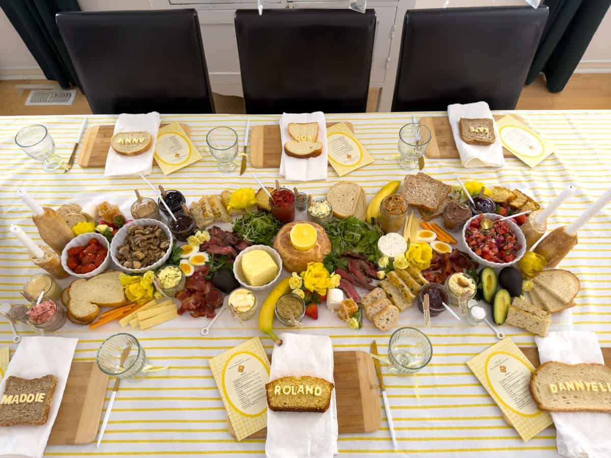 Butter themed toast party dining table set for six with yellow striped tablecloth, compound butter grazing spread down the center, lit taper candles in baguette holders, butter candle in sourdough boule, name bread place settings, and gallery wall of botanical prints visible in background.