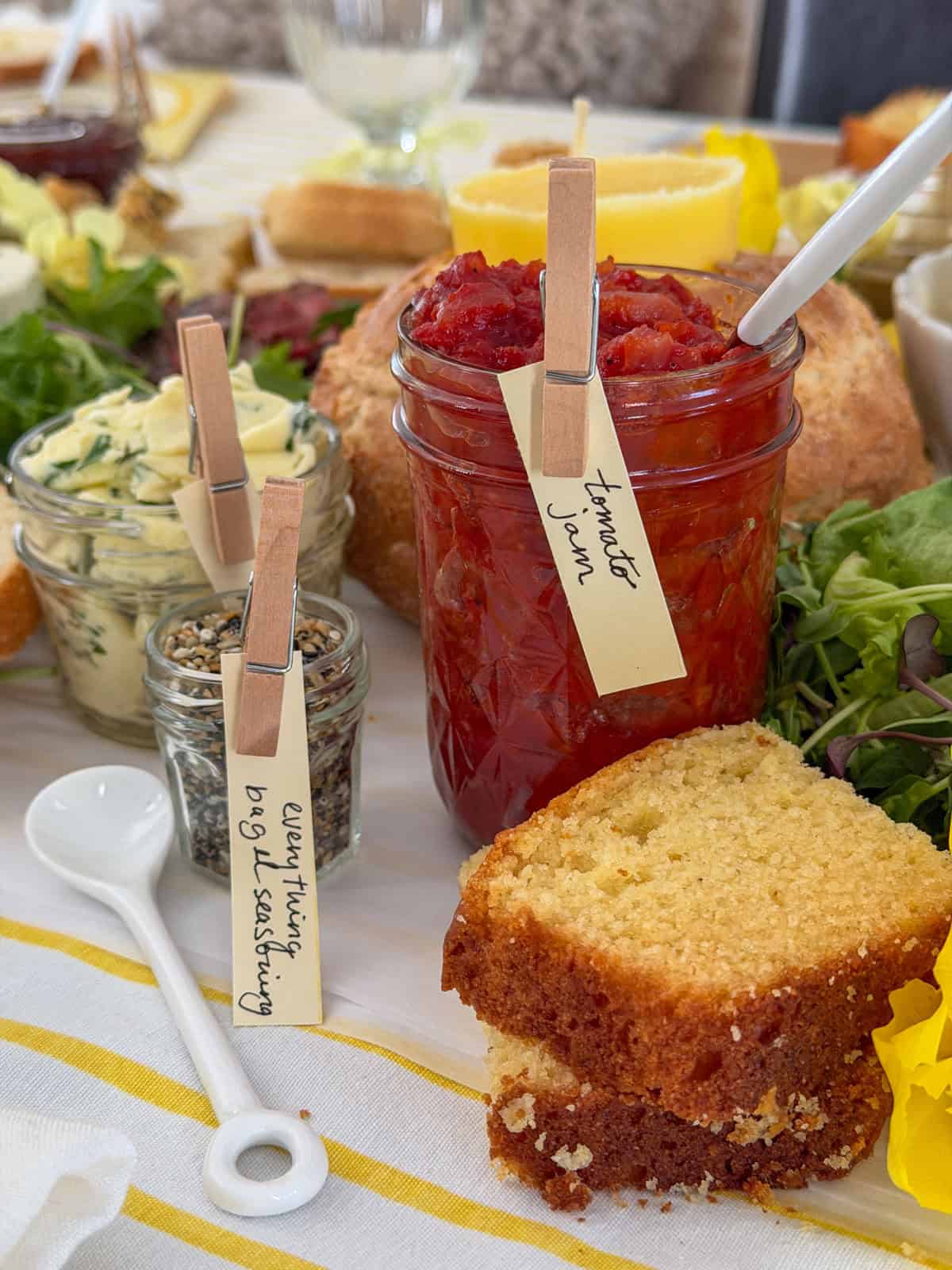 Close up of tomato jam mason jar with handwritten clothespin label beside a jar of everything bagel seasoning on a white parchment grazing surface with garlic herb butter jar, slice of cornbread, yellow tulip, and butter candle in sourdough boule visible in background.